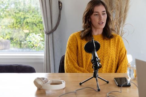 Woman recording cozy home podcast wearing mustard sweater at wooden table