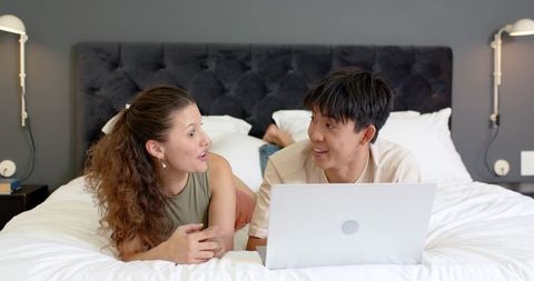 Young asian man and african american woman relaxing on bed using laptop and chatting