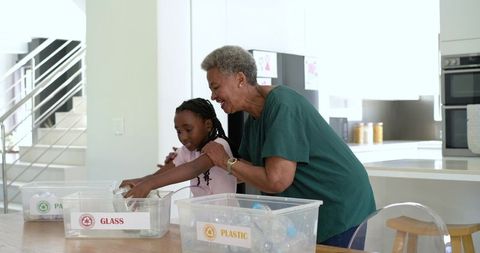 Senior african woman mentoring girl sorting glass and plastic recyclables in sunlit kitchen