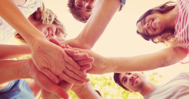A diverse group of young people is stacking hands together in a gesture of teamwork and unity, with copy space. Their smiling faces and the warm, sunlit setting convey a sense of friendship and cooperation.