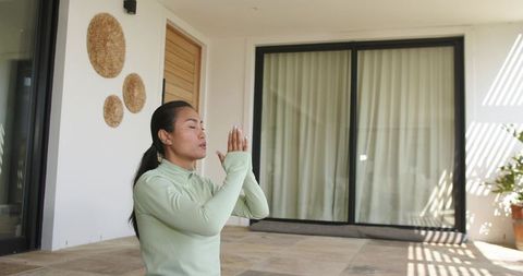 Asian woman practicing meditation on sunlit terrace kneeling in green active top peaceful