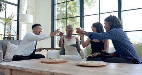 Diverse friends raising wine glasses, sharing pizza and celebrating in modern living room