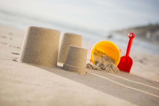 Sand castles standing proudly on a sunny beach alongside a colorful yellow bucket and red spade. Ideal for themes of summer vacations, family fun, and beach activities. Perfect for use in travel brochures, family holiday advertisements, and outdoor play promotions.