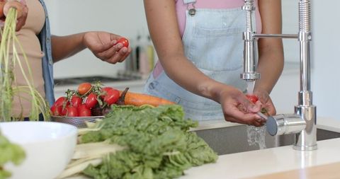 Mother and teen daughter washing fresh tomatoes and greens together at kitchen sink home cooking