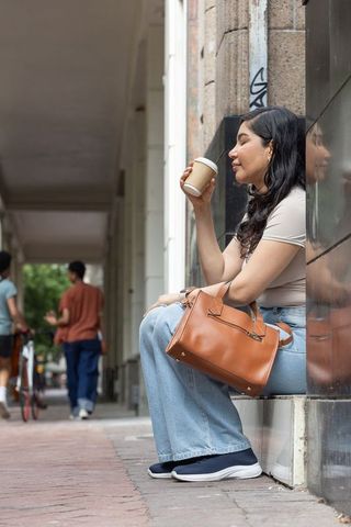 Sipping coffee on urban arcade ledge, casual woman resting with leather handbag