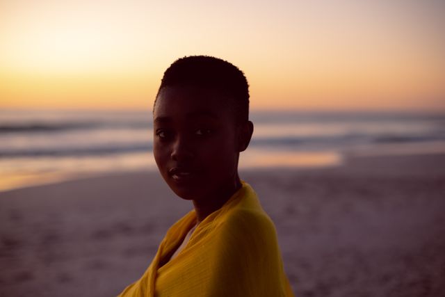 This image captures a young woman wrapped in a yellow scarf standing on a beach at sunset. The serene and tranquil setting with the ocean in the background makes it perfect for use in travel blogs, fashion magazines, wellness articles, and advertisements promoting relaxation and beach destinations.