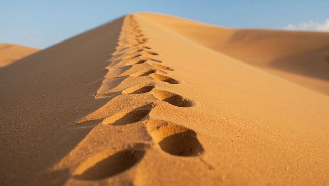 Footprints climbing dune crest in vast golden desert under clear blue sky