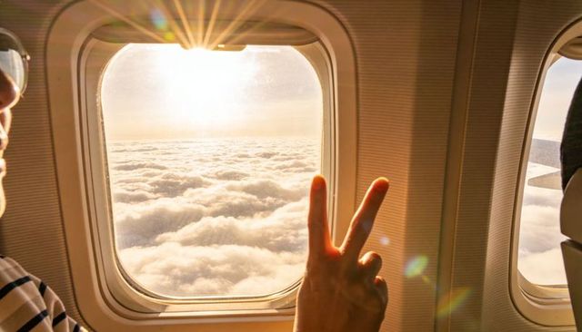 Passenger making peace sign framing golden clouds through airplane window at sunrise