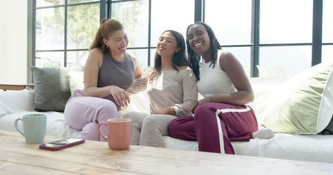 Three friends relaxing on sunlit sofa sharing coffee and laughter in cozy living room