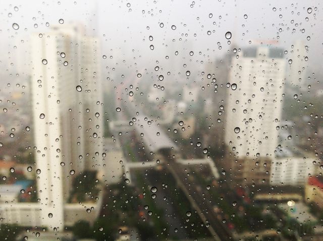 Raindrops on window creating an artistic view of an urban cityscape with high-rise buildings in background. Suitable for illustrating weather conditions, urban life, or themes of melancholy and reflection. Ideal for websites, blogs, and advertisements related to weather, real estate, or urban living.