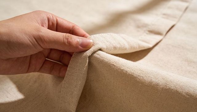 Left hand pinching folded beige linen fabric showing seam texture and woven detail