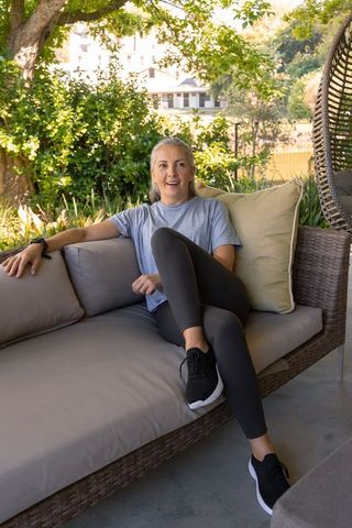 Senior woman relaxing on woven patio sectional wearing smartwatch and sneakers in shaded backyard