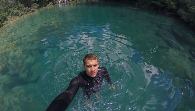 Self-filming man wading in turquoise lake with waterfall and forest reflections