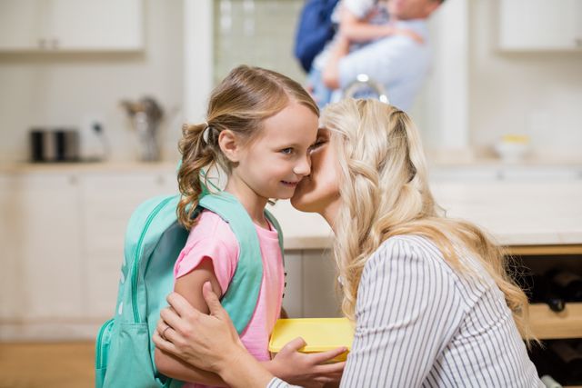 Mother kissing daughter on forehead in kitchen before school. Daughter wearing backpack, mother holding lunchbox. Ideal for use in family, parenting, education, and morning routine themes.