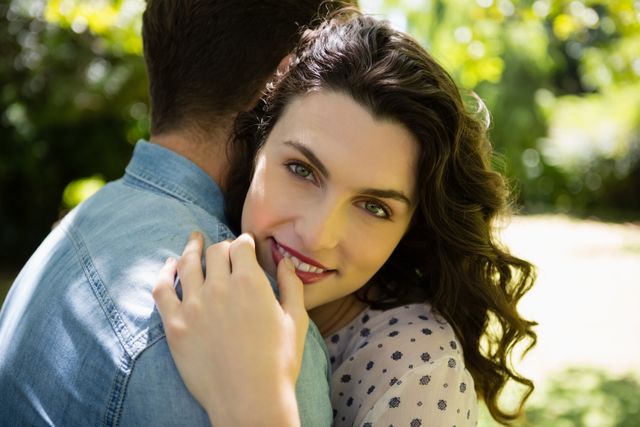 Couple embracing in a lush garden on a sunny day. Woman smiling warmly while holding her partner. Ideal for use in relationship blogs, romantic greeting cards, love-themed advertisements, and lifestyle magazines.