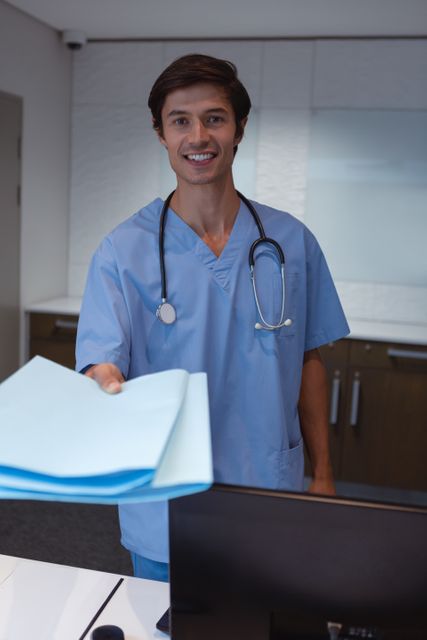 Male surgeon in blue scrubs holding documents and smiling at hospital desk. Ideal for healthcare, medical services, hospital administration, and patient care themes.