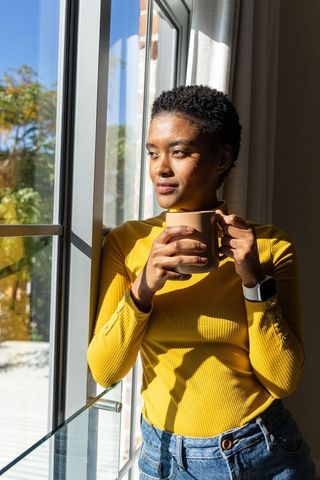 African american woman sipping coffee and gazing out sunlit window in casual morning routine