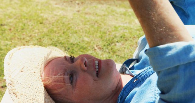 Elderly woman lying on grass, wearing straw hat and smiling. Perfect for themes related to relaxation, outdoor activities, senior lifestyle, enjoying nature, and leisure time. Can be used in health and wellness articles, retirement plans, vacation brochures, or advertisements promoting relaxed, happy living.