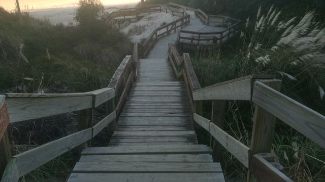 Wooden pathway leads down to the beach during sunset, creating a tranquil and serene atmosphere. Sand dunes and surrounding vegetation add to the natural beauty. Ideal for travel articles, website backgrounds, posters, and nature blogs about beach destinations and outdoor relaxation.