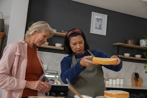 Senior women assembling cake layers in home kitchen, baking together and inspecting sponge