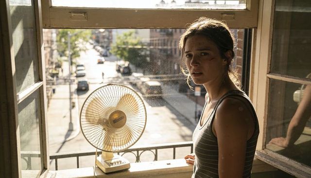 Bruised woman standing at open apartment window with fan and city street at golden hour