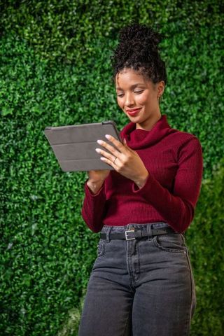 African american woman using tablet outdoors against hedge wall in burgundy turtleneck