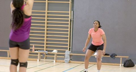 Diverse female teammates practicing volleyball drill on gym court wearing pink shirts