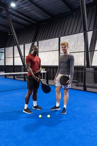 Diverse male friends standing on blue indoor padel court holding rackets and balls