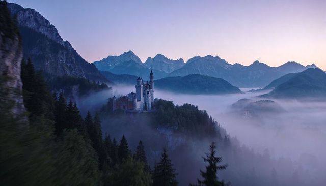 Perching fairytale castle looming above mist-filled pine valley at dawn with alpine peaks