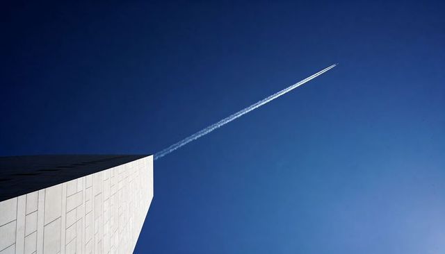 Minimalist diagonal monument corner, stone facade meeting jet contrail over deep blue sky