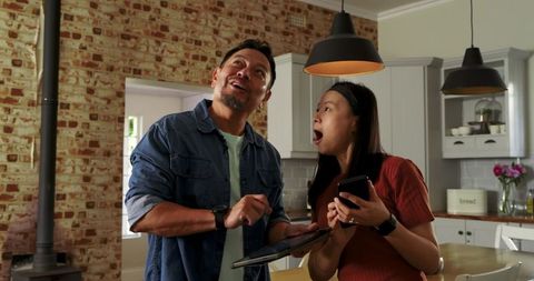 Asian couple reacting excitedly while using tablet and smartphone in farmhouse kitchen