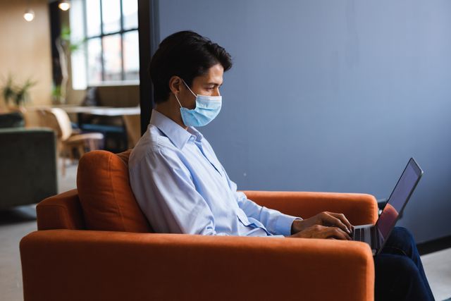 Biracial businessman wearing face mask using laptop sitting in armchair. health and hygiene in workplace during coronavirus covid 19 pandemic.