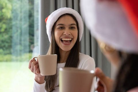 Young woman wearing santa hat laughing and holding mug by window, cozy holiday moment