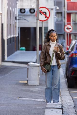African american woman walking narrow urban street holding coffee cup in brown jacket