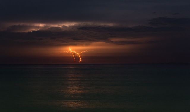 Dramatic lightning strike illuminating dark clouds over calm ocean. Ideal for concepts related to nature's power, energy, environmental themes, maritime storytelling, or backgrounds in weather-related television shows.