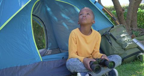 Young african american boy sitting at tent door holding binoculars gazing upward