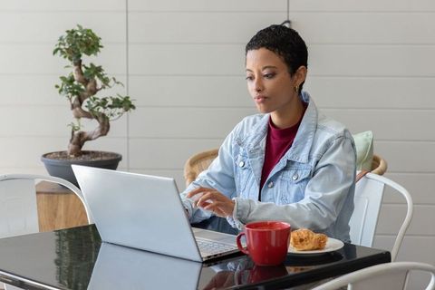 African american woman working on laptop in modern cafe with red mug and croissant