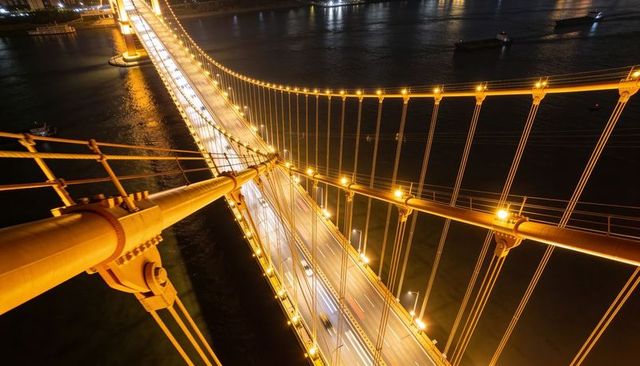 Aerial night view revealing golden suspension bridge cables and light trails over river
