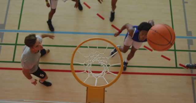 Basketball players are competing to score a goal, seen from above the hoop. One player is shooting the ball while others are preparing for the rebound. Could be used in sports promotions, basketball training materials, or team marketing content.