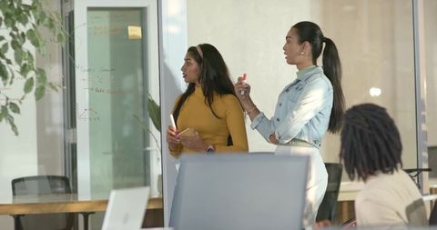 Two women collaborating at glass wall brainstorming with marker and notebook, colleague watching