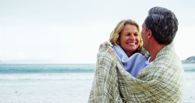 Happy senior couple hugging under a blanket on a beach during autumn. They are smiling, capturing a warm and affectionate moment. Ideal for promoting romance in older adulthood, senior lifestyle, outdoor and beach activities, health and wellness for seniors, and travel advertisements.