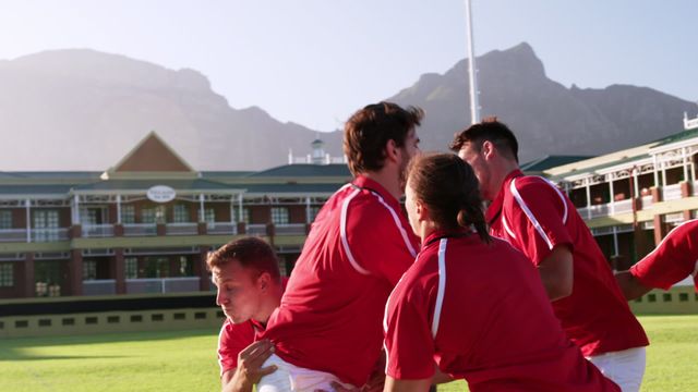 Rugby players in red jerseys lift a teammate to catch the ball during a game in a sunny stadium with mountains in the background. Useful for sporting events, teamwork demonstrations, athletic promotions, or rugby-related content.