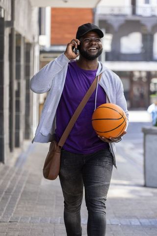 African american man walking city street holding basketball and talking on phone smiling
