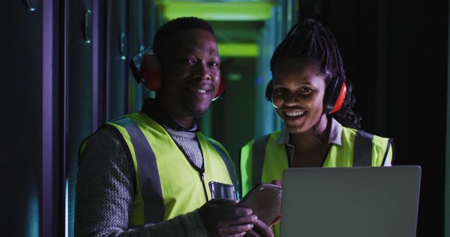 African American technicians in safety vests working in a server room data center. One holds a laptop, while both wear ear protection. Suitable for illustrating teamwork in technology and IT infrastructure.