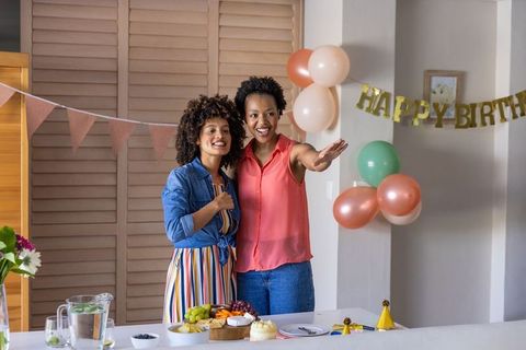 Two diverse women celebrating birthday at home with cake, pastel balloons, brunch table