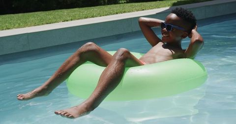 Teenager relaxing on neon green inner tube floating in backyard pool wearing sunglasses