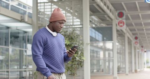 African american man walking transit terminal using smartphone pulling luggage headset