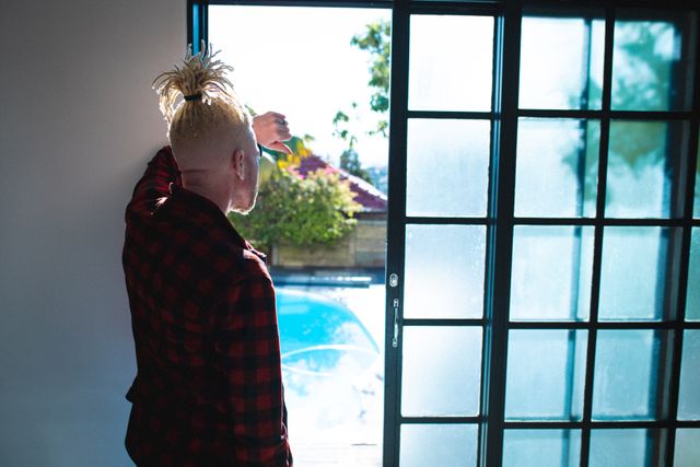 Man with dreadlocks standing by window, looking outside. Ideal for themes of contemplation, relaxation, leisure time, and lifestyle. Can be used in articles about mental health, personal reflection, or home life.