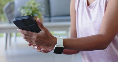 Woman holding smartphone with rugged case and white smartwatch, tapping screen outdoors