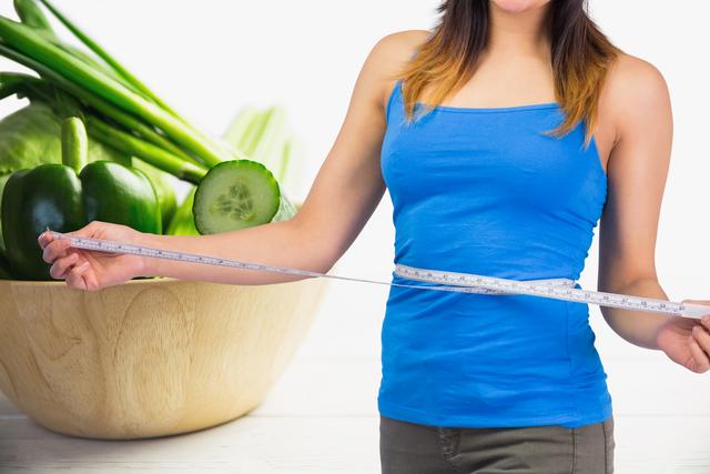 Midsection of a woman wearing a blue tank top measuring her waist, emphasizing her commitment to a healthy lifestyle. Fresh green vegetables including cucumbers and bell peppers in a wooden bowl on a white background reinforce the message of healthy eating and wellness. Ideal for use in articles, advertisements, and blogs about dieting, nutrition, weight loss, fitness, and promoting healthy lifestyle choices.